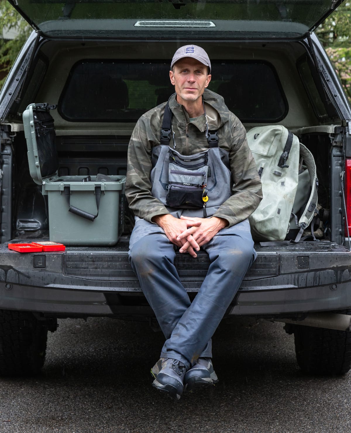 A man sits on the tailgate of a truck, facing the camera, with fly fishing gear on either side of him, ready to give a briefing to a beginner before setting out.