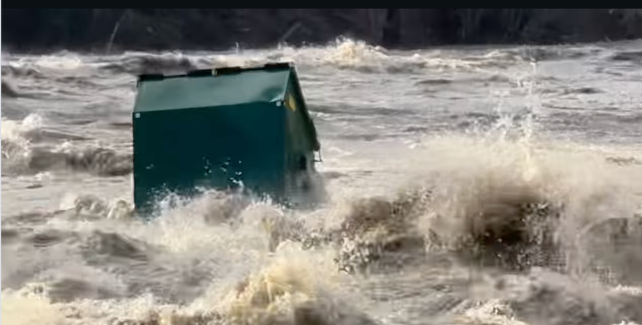 A big green Dumpster floating down a flooded, turbulent, muddy river, with splashing all around it. 