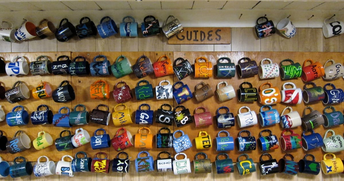 A wall labeled 'GUIDES' holds approximately 100 handmade coffee mugs arranged in horizontal rows. Each mug is hung by its handle and features a name, forming a colorful mosaic.
