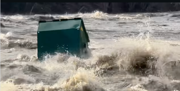 A big green Dumpster floating down a flooded, turbulent, muddy river, with splashing all around it. 