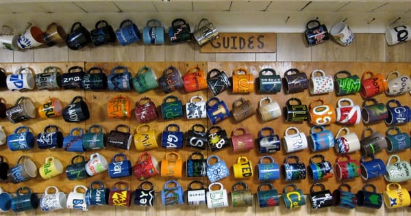 A wall labeled 'GUIDES' holds approximately 100 handmade coffee mugs arranged in horizontal rows. Each mug is hung by its handle and features a name, forming a colorful mosaic.